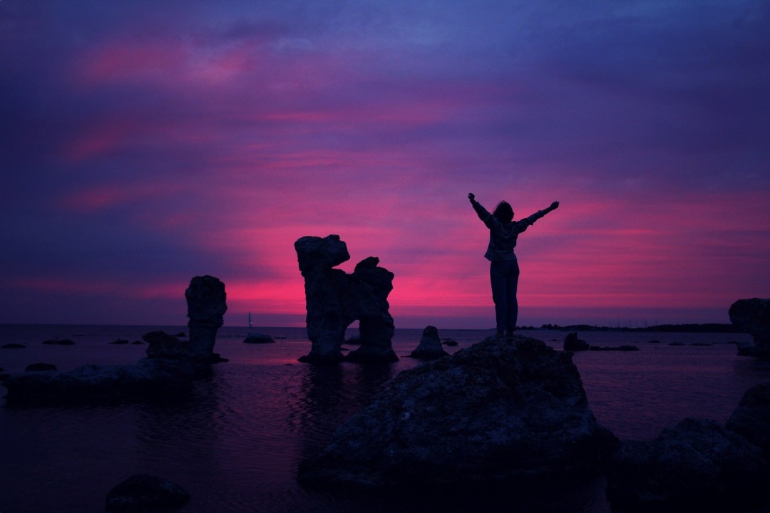 Woman standing on a rock with arms raised overlooking pink and purple sky